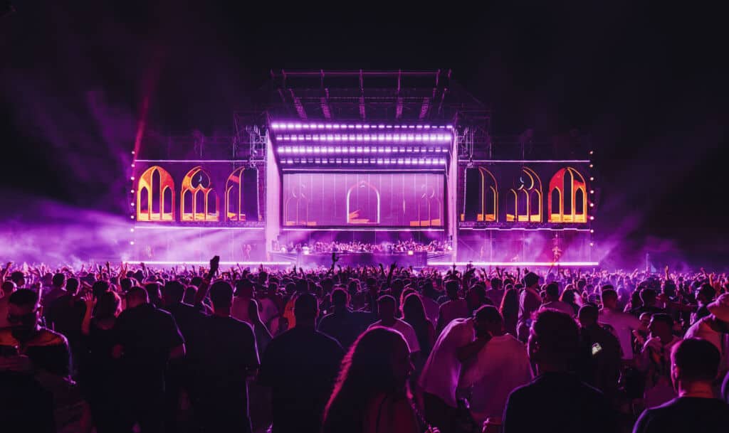 Un palcoscenico illuminato con luci neon rosa e arancioni, atmosfera da festival musicale notturno con pubblico entusiasta.